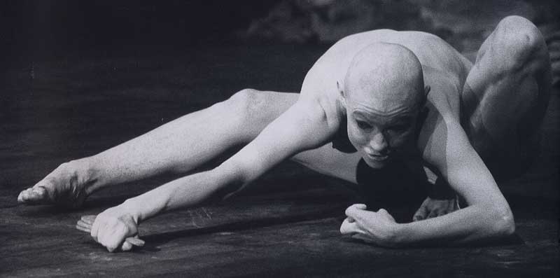 Dançarino de Butô com o corpo pintado de branco em uma pose contorcida no chão.