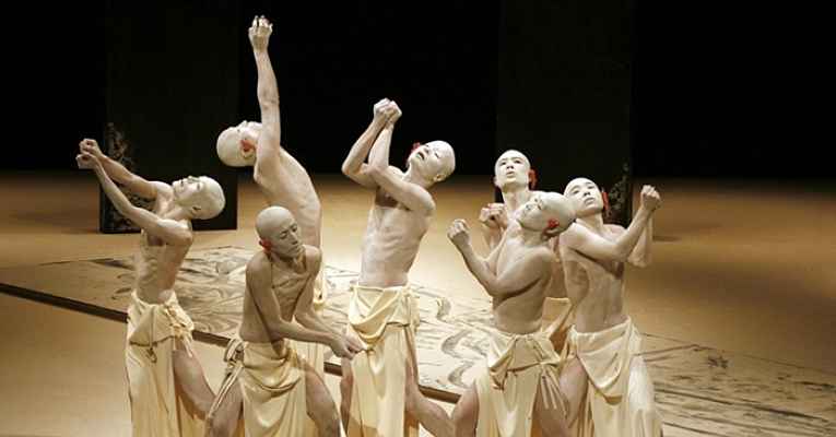Grupo de dançarinos de Butô com corpos brancos e cabeças raspadas em uma formação ao ar livre.