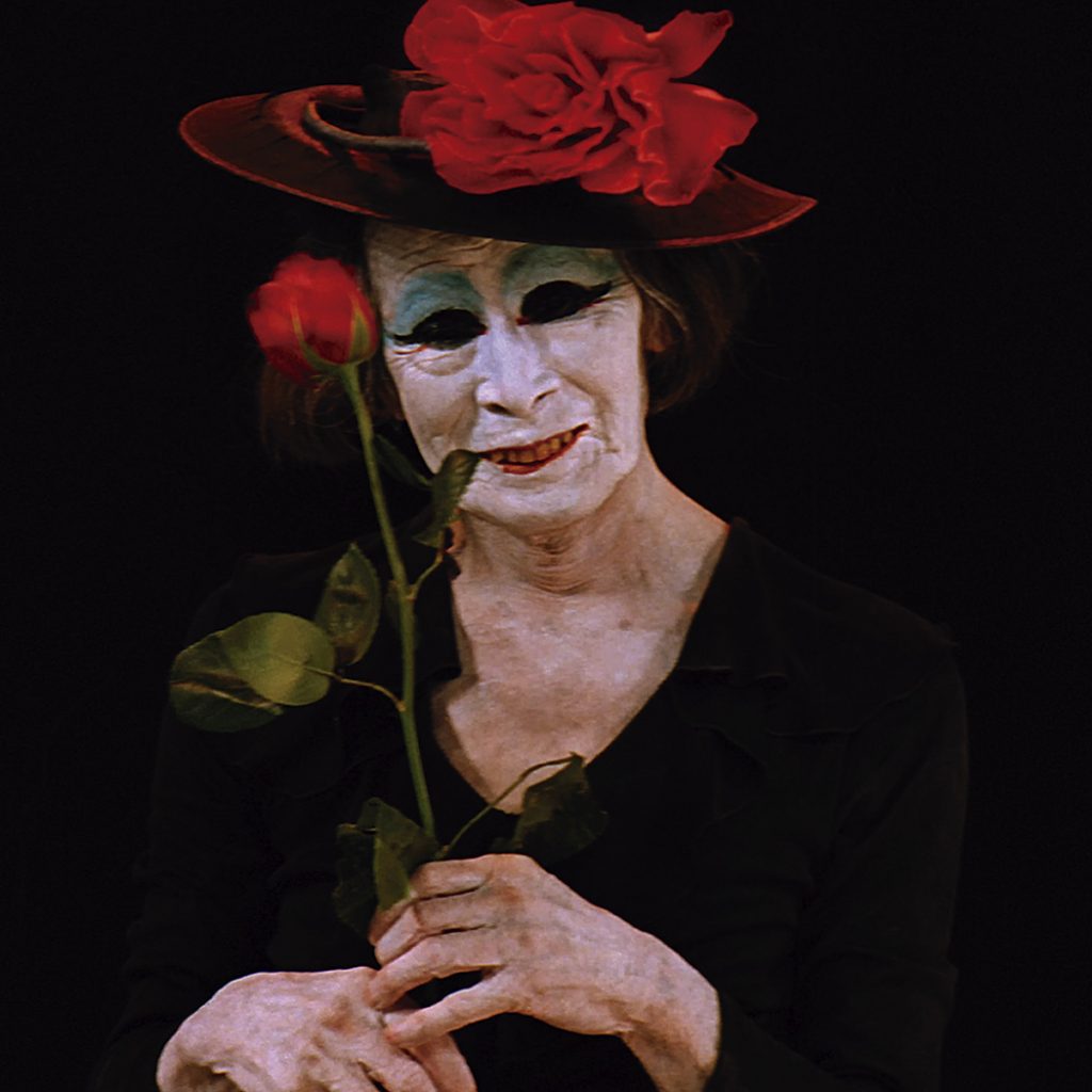 Kazuo Ohno, um homem idoso de rosto enrugado e pintado de branco, com uma expressão intensa e olhos fechados, vestindo um chapéu.